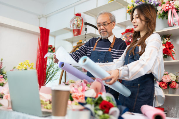 小企业主使用笔记本电脑和看着相机在花店。亚洲年长男性花店老板与年轻漂亮的女员工讨论花束布置和装饰业务图片下载