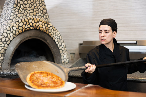 披萨店的厨房里，身穿黑色制服的女厨师正在准备披萨，并将其从烤箱中取出图片下载