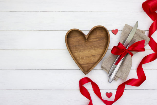 情人节的场所设置。木制心形盘子，刀叉配红蝴蝶结，白色木板上系红丝带图片下载