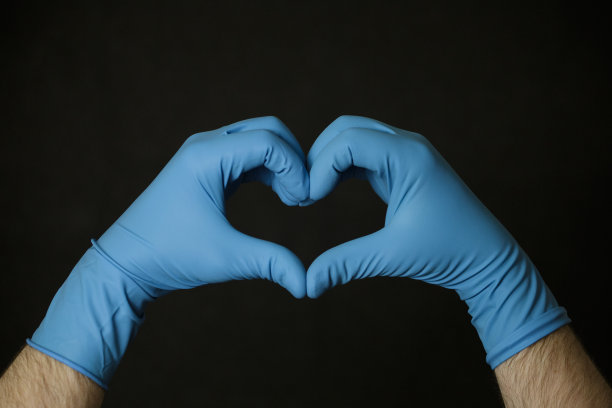 一个戴着蓝色橡胶手套的人在黑色背景上用自己的手做了一个心，选择性的聚焦。关心和保护的概念。一位戴着橡胶手套的心脏病专家展示了心脏。图片下载