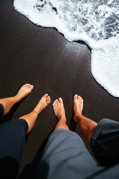 一对夫妇的脚在黑沙滩上的软泡沫波的高角度视图图片下载