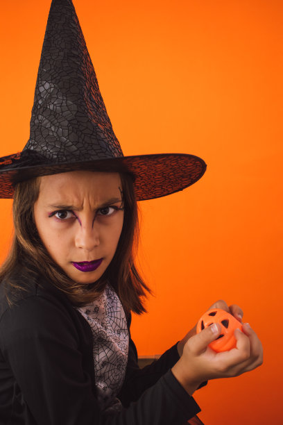 一个孩子穿着女巫服装，手里拿着一个南瓜，橙色背景的垂直镜头图片下载