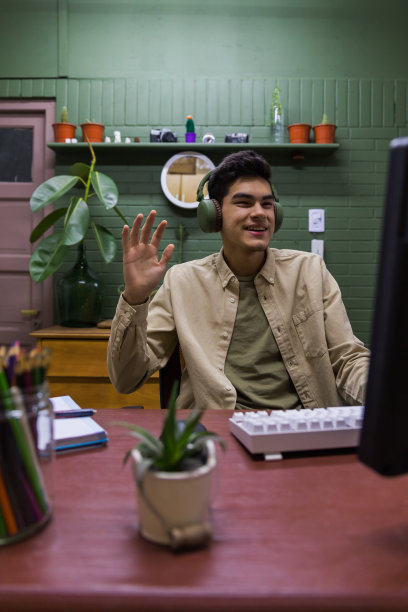 一名西班牙裔男性手持耳机坐在电脑前挥手图片下载
