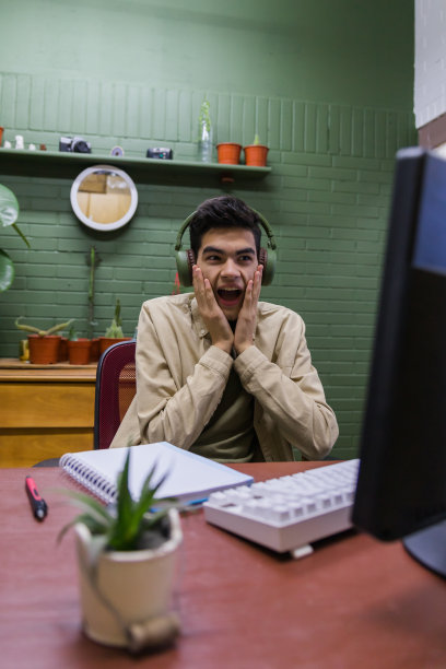 垂直拍摄一位惊讶的西班牙裔男性，戴着耳机坐在电脑前的桌子旁图片下载
