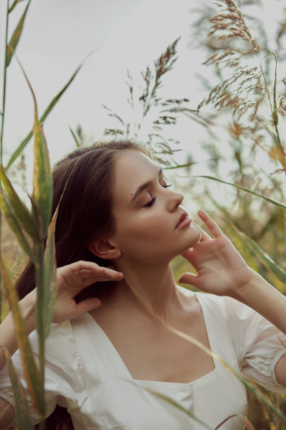 身着白色夏装的美丽女子正坐在乡间的草丛中。夕阳下浪漫女孩的肖像，温暖的太阳，自然美丽的女人图片下载