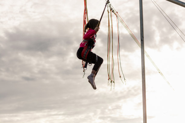女孩正在蹦床上跳。一个带着保险和可拉伸橡皮筋的孩子悬在空中。快乐童年的概念和游乐园的游戏。图片下载