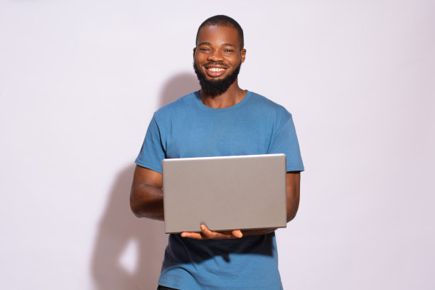 一个穿着蓝色t恤的黑人在白色背景上微笑着拿着笔记本电脑。图片下载