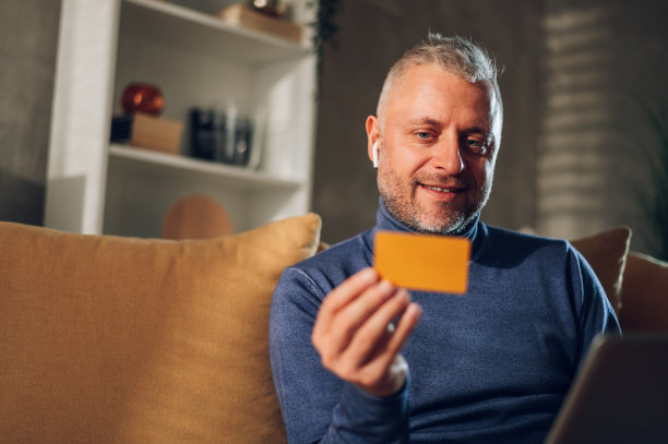 一名中年男子坐在家里的沙发上使用笔记本电脑和信用卡图片下载