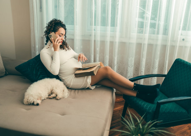年轻的孕妇在她公寓的沙发上看书，她的宠物狗和她在一起图片下载