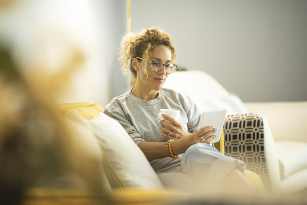 微笑的中年白人女性坐在客厅的沙发上浏览平板电脑上的无线网络，快乐的现代漂亮女性在家里的沙发上放松使用平板设备，黄色，科技概念图片下载