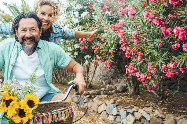 年轻成熟的夫妇一起享受户外休闲活动，有乐趣和欢笑。一男一女在外面骑自行车，背景是花。春天健康可持续生活方式图片下载