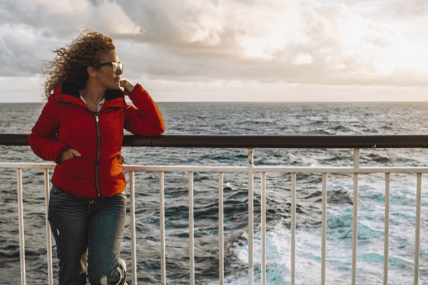 一位穿着红色夹克的女子在夕阳下欣赏着海面。背景是美丽的天空。女性乘坐轮渡、游船旅行的概念。游客享受交通图片下载