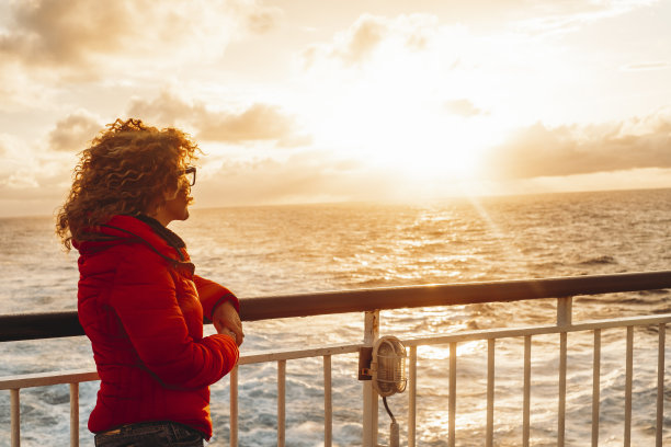旅行是人们生活方式的概念。一名女子站在渡轮巡游甲板上，欣赏海洋上的夕阳，背景是水。旅行者和漫游癖。地平线与天空的背景图片下载