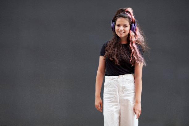 年轻快乐的女孩带着粉红色的长发，戴着耳机听着音乐，微笑着看着深灰色背景上的相机图片下载