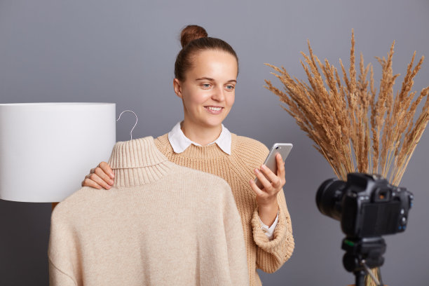 照片中，穿着休闲服装的女造型师面带微笑，站在灰色的墙上，背景是灯和干花，用相机和手机录制视频。图片下载