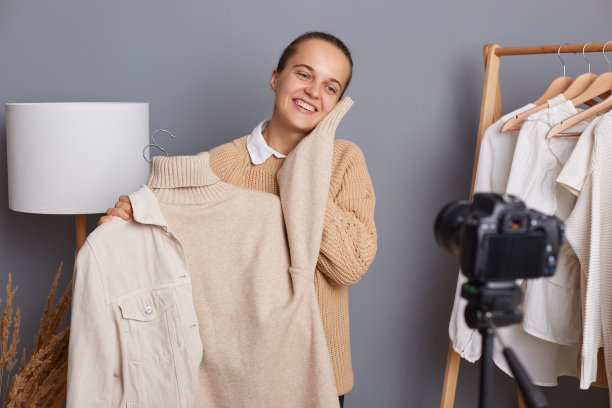 可爱迷人的满意微笑的女人穿着米色的套头衫，显示温暖柔软的毛衣，为她的vlog创建内容，录制视频，站在灰色的墙上，衣服挂在架子上。图片下载
