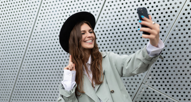 一名戴着黑色帽子、穿着灰色外套的时尚年轻女子在一座办公楼的非铝制立面背景下自拍图片下载