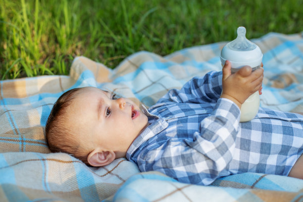 可爱的宝宝在野餐时躺在毯子上喝瓶子里的牛奶。婴儿的营养。图片下载