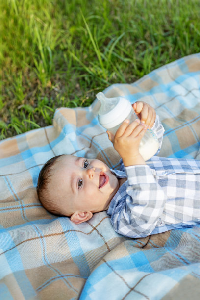 微笑。可爱的宝宝在野餐时躺在毯子上喝瓶子里的牛奶。婴儿的营养。图片下载