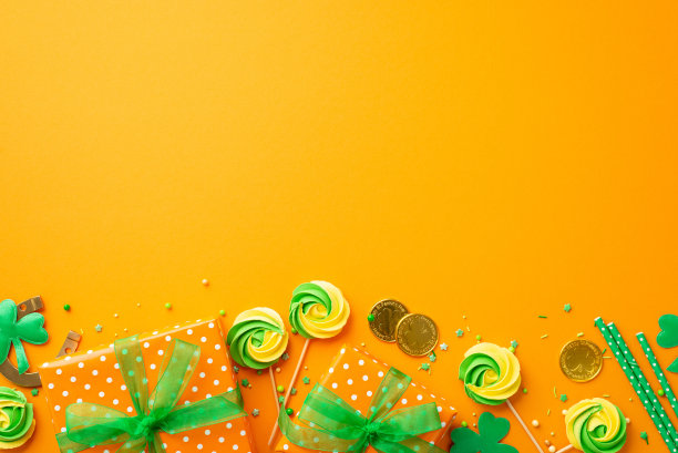 圣帕特里克节的概念。俯视图照片的礼物盒与绿色丝带蝴蝶结蛋白糖棒棒糖洒金币马蹄铁和三叶草孤立的橙色背景与空白图片下载