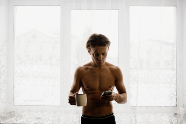 运动男子与一个赤裸的躯干喝杯在早上靠窗。图片下载