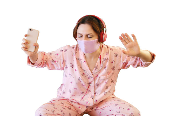 一名妇女在感染新冠流感病毒期间坐在床上听音乐和跳舞，白色背景隔离。穿着睡衣，戴着耳机和防护面具的女人图片下载