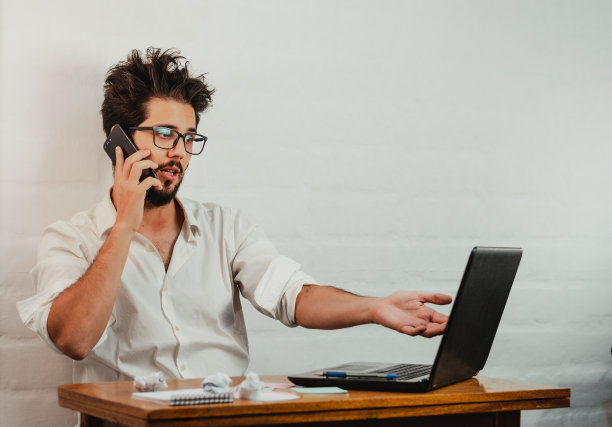 年轻的白人男性在办公室打电话时看着笔记本电脑并打手势图片下载