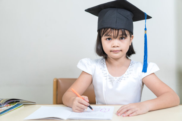 一个亚裔小女孩的肖像，戴着学士帽，坐在桌子旁，在她的抄写本上写字图片下载