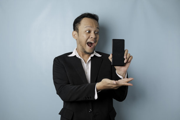 惊讶的亚洲商人穿着黑色西装，拿着他的智能手机，屏幕上显示出被蓝色背景隔离的拷贝空间图片下载