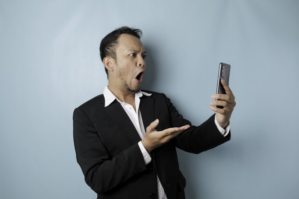 惊讶的亚洲商人穿着黑色西装拿着他的智能手机，被蓝色背景隔离图片下载