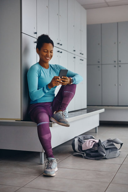 快乐的黑人女运动员在健身房的更衣室用手机发短信。图片下载