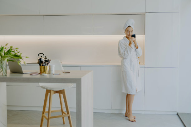 完整的镜头，年轻美丽的女子穿着白色浴袍，涂面霜，照镜子，照顾她的皮肤，在厨房家具附近摆姿势，赤脚站立。人们美容面部护理图片下载