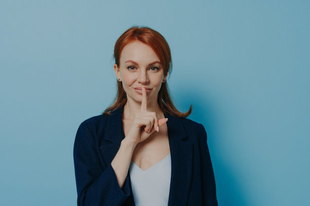 年轻的红发女子要求保持沉默或不透露秘密，孤立在蓝色背景上图片下载