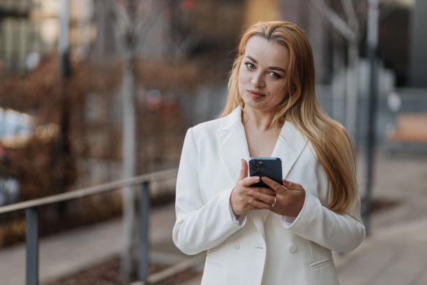 金发时尚的女人在超大的白色夹克上使用手机在现代城市的背景图片下载