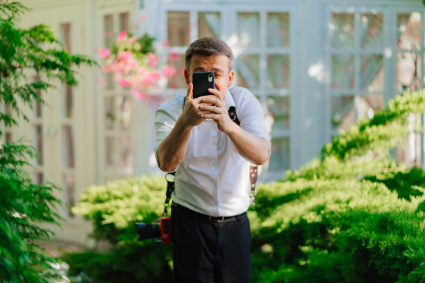 一名身穿白衬衫的男子在公园里用智能手机拍照。图片下载