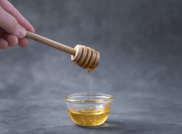 在一个深色背景的特写中，蜂蜜从一根木棍流进一个玻璃碗里。健康有机食品的概念。图片下载