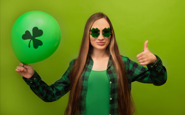 年轻女子在绿色格子衬衫三叶草形状的眼镜显示大拇指或推荐你和持有气球与三叶草打印孤立在绿色背景。  圣帕特里克节。图片下载