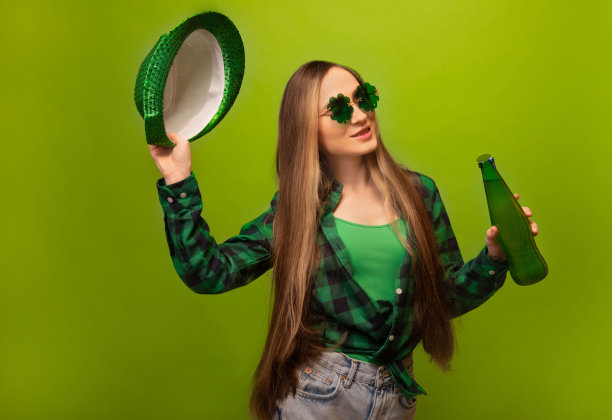 快乐的微笑年轻女子在绿色格子衬衫三叶草形状的眼镜持有绿色啤酒瓶和聚会帽孤立在绿色背景。  圣帕特里克节庆祝概念。图片下载
