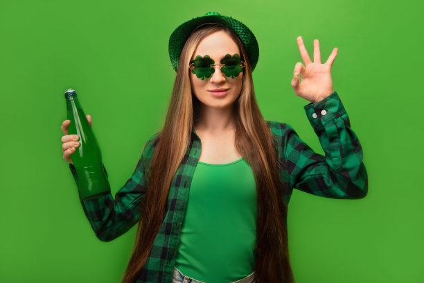 时尚快乐的女孩显示ok手势，穿着格子衬衫，派对绿色帽子与三叶草形状的眼镜，拿着绿色啤酒瓶在彩色绿色背景。

圣帕特里克节。图片下载