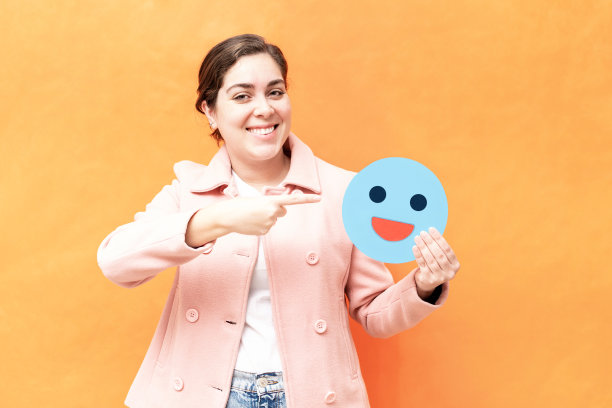 快乐的白人女孩拿着一张蓝色表情符号的纸，孤立在橙色背景上图片下载