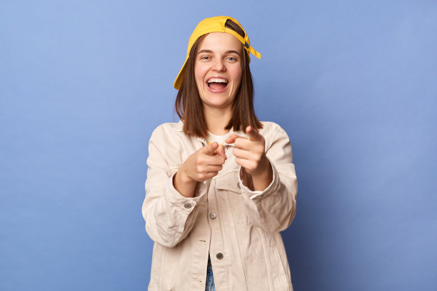 可爱迷人的积极棕色头发的少年女孩戴棒球帽和夹克的照片在蓝色背景上孤立地摆姿势，指着相机，选择你。图片下载