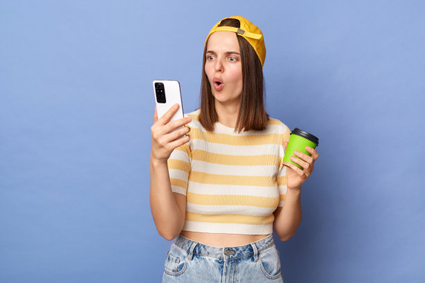 照片中的惊讶震惊的少女戴着棒球帽和休闲t恤，喝着咖啡和使用手机，表达震惊，孤立地站在蓝色背景上。图片下载