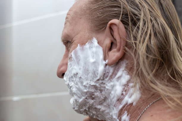 一个脸上涂着厚厚的剃须泡沫的成熟男人在浴室里刮胡子。护肤卫生图片下载