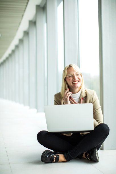 年轻的金发女商人在笔记本电脑上工作，用手机交谈图片下载