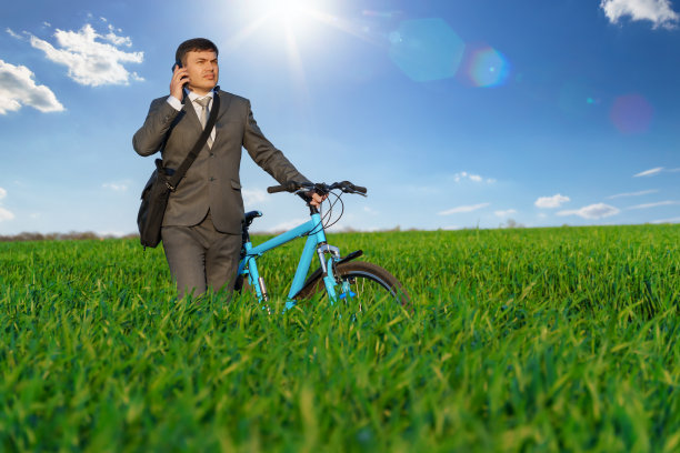 一个商人骑着自行车在绿油油的草地上，穿着西装，他在打一个电话，美丽的大自然在春天，自由职业者的商业概念图片下载
