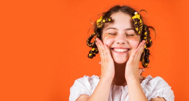 造型技巧。青少年的爱好。小女孩卷发使用卷发器橙色背景。理发师沙龙。女性美容套路。可爱的儿童发型。戴着卷发棒的女儿在笑图片下载