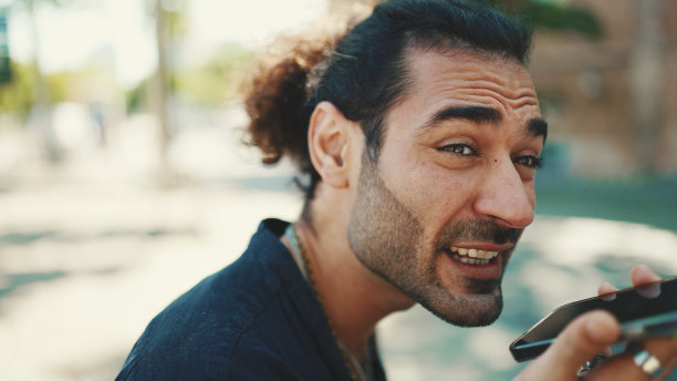 迷人的意大利男子与马尾和胡茬正在使用手机坐在长凳上在老建筑背景。型男写留言，用智能手机看照片图片下载