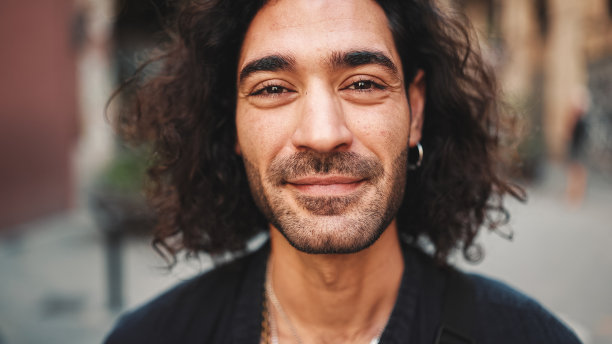 年轻迷人的意大利男子长卷发和胡茬看着相机和微笑在老建筑的背景。时髦男人，耳朵上戴着耳环，戴着很多链子图片下载