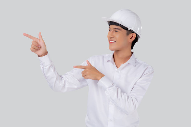 肖像年轻的亚洲男子是工程师戴着头盔站着，微笑着自信孤立的白色背景，男性是建筑师或承包商，展示或展示一些工业概念。图片下载