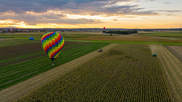 鸟瞰图热气球站在草地上，旁边有多个农田，联合收割机正在割玉米，拖拉机和拖车停在田地旁边图片下载